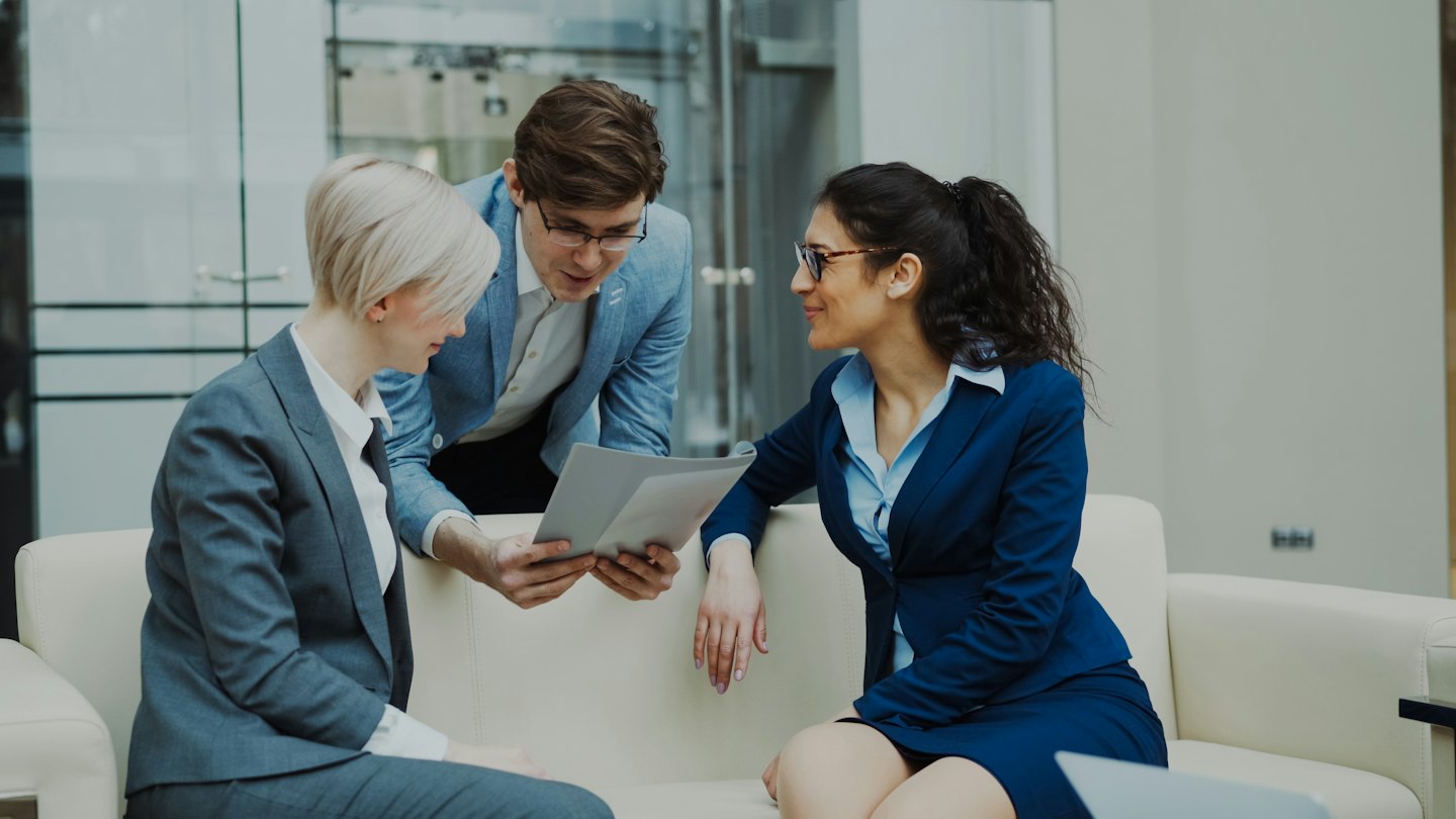 Three colleagues discuss business documents in a modern office lounge.